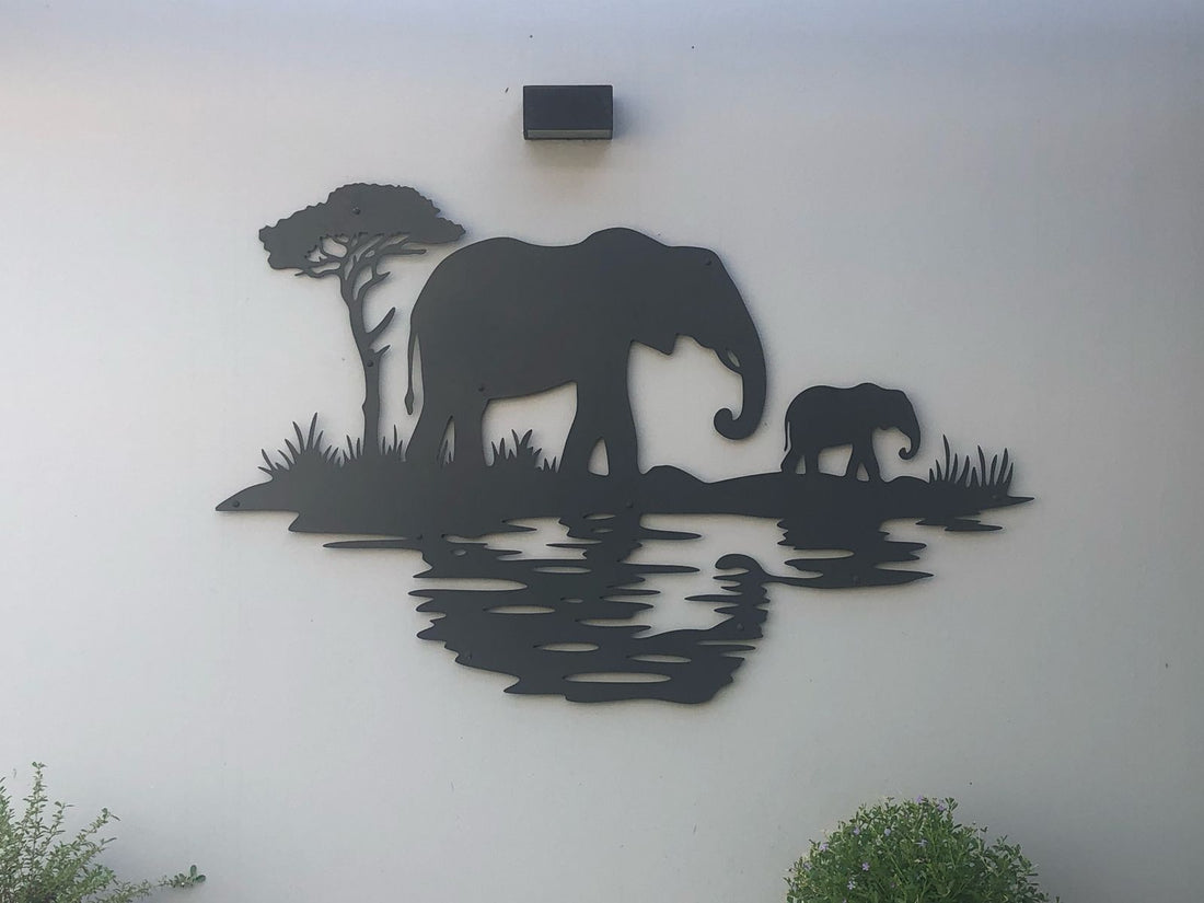 A black metal wall art design depicting an adult elephant and a calf standing near the edge of a reflective water body, accompanied by a solitary tree in the background.