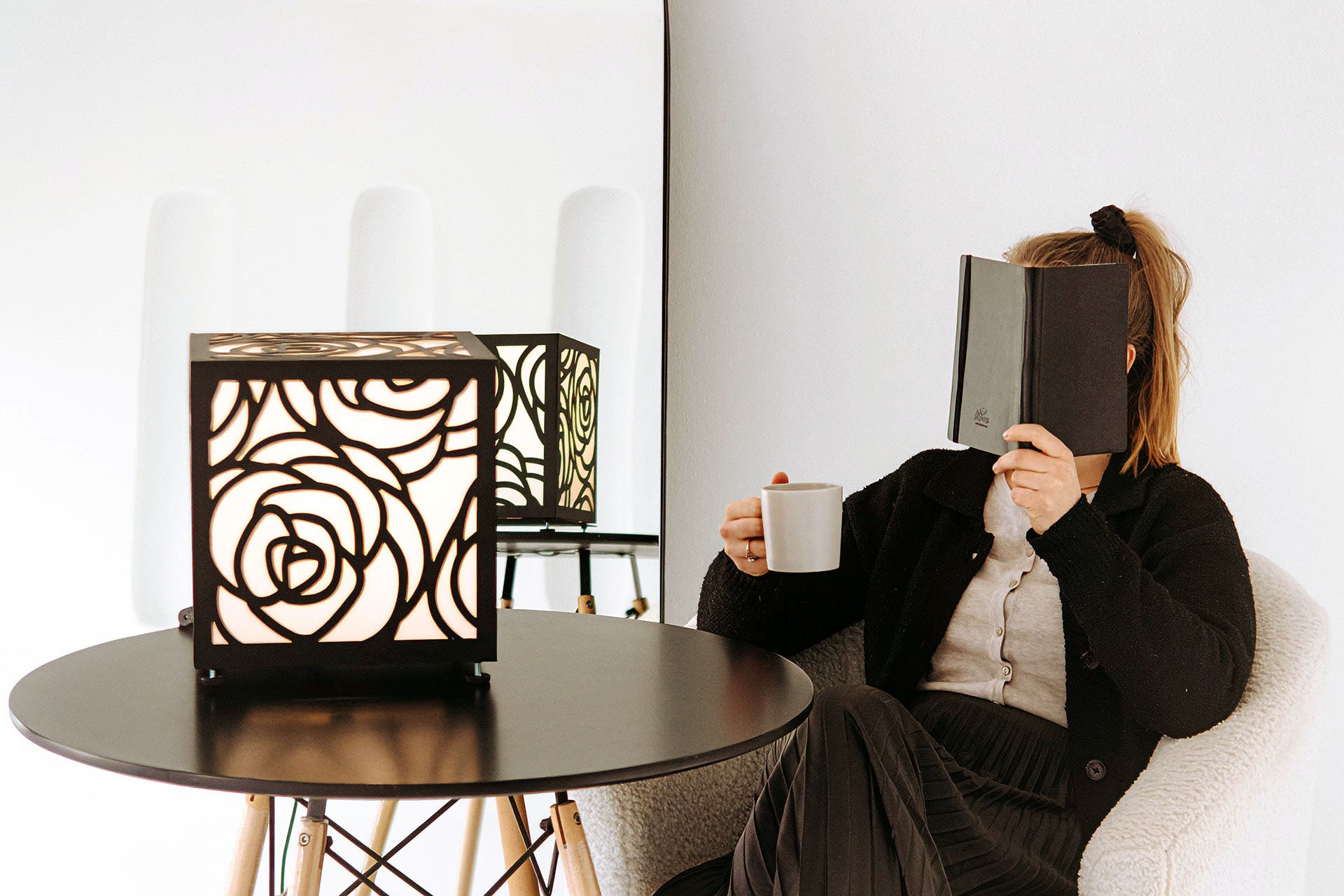 Person sitting on a white chair reading a book with a decorative table lamp in the background.